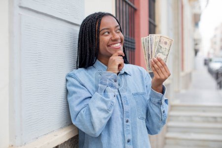 Foto de Mujer afro negro sonriendo con una expresión feliz y segura con la mano en la barbilla. concepto de billetes en dólares - Imagen libre de derechos