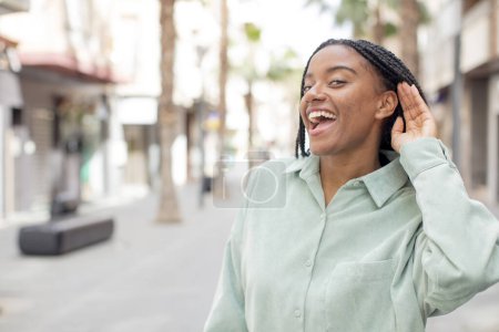 Téléchargez les photos : Afro jolie femme noire regardant heureux, écoutant, essayant d'entendre une conversation secrète ou des commérages, écoutant - en image libre de droit