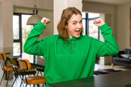 Photo for Pretty caucasian woman feeling happy, satisfied and powerful, flexing fit and muscular biceps, looking strong after the gym. home interior concept - Royalty Free Image