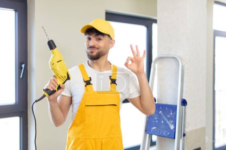 Photo for Young handsome man feeling happy, showing approval with okay gesture. handyman and drill concept - Royalty Free Image
