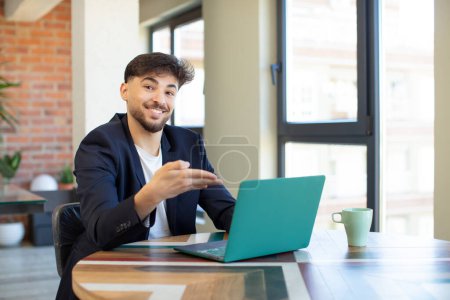Foto de Joven hombre guapo sonriendo alegremente, sintiéndose feliz y mostrando un concepto. concepto freelance con portátil - Imagen libre de derechos