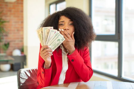 Foto de Mujer negra bastante afro que cubre la boca con una mano y la expresión impactada o sorprendida. concepto de billetes en dólares - Imagen libre de derechos