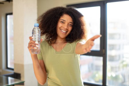 Téléchargez les photos : Jolie afro femme noire souriant joyeusement et offrant ou montrant un concept. concept de bouteille d'eau - en image libre de droit