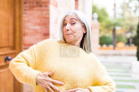 Photo for Senior retired pretty white hair woman with a goofy, crazy and surprised expression, feeling stuffed, fat and full of food - Royalty Free Image