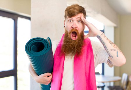 Foto de Hombre de pelo rojo que se ve feliz, sorprendido y sorprendido con un mate de yoga. concepto de fitness - Imagen libre de derechos