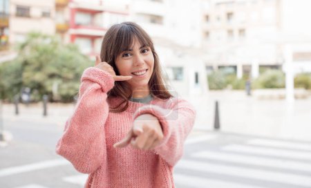 Photo for Pretty young woman smiling cheerfully and pointing to camera while making a call you later gesture, talking on phone - Royalty Free Image