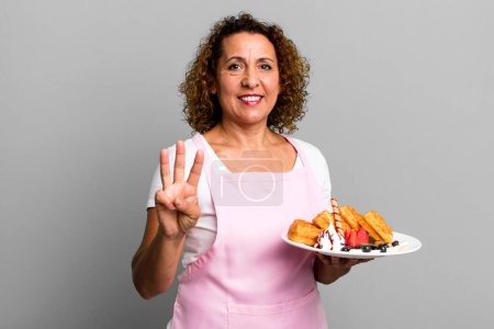 Foto de Bastante mujer de mediana edad sonriendo y buscando amigable, mostrando el número tres. concepto de gofres caseros - Imagen libre de derechos
