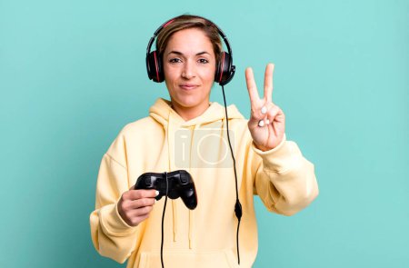 Foto de Bonita mujer rubia sonriendo y buscando amigable, mostrando el número dos. gamer con auriculares y un controlador - Imagen libre de derechos