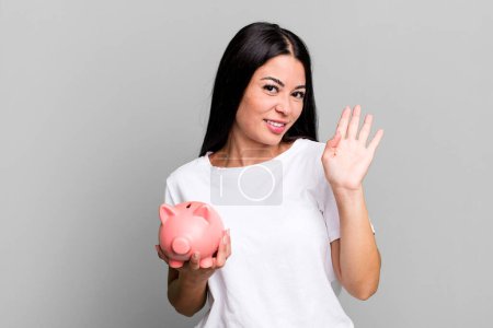 Téléchargez les photos : Hispanic pretty woman smiling happily, waving hand, welcoming and greeting you with a piggy bank - en image libre de droit