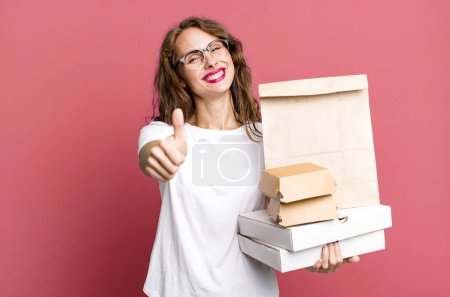 Foto de Mujer bonita hispana sintiéndose orgullosa, sonriendo positivamente con los pulgares hacia arriba. quitar concepto de paquetes de comida rápida - Imagen libre de derechos