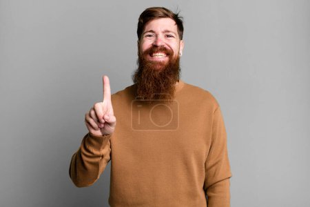 Foto de Barba larga y pelo rojo hombre sonriendo y buscando amigable, mostrando el número uno - Imagen libre de derechos