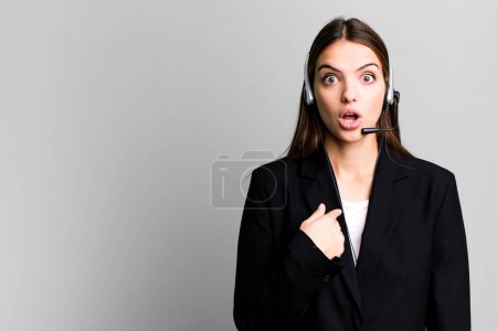 Foto de Joven bonita mujer mirando sorprendido y sorprendido con la boca abierta, apuntando a sí mismo. concepto de agente de telemarketing - Imagen libre de derechos