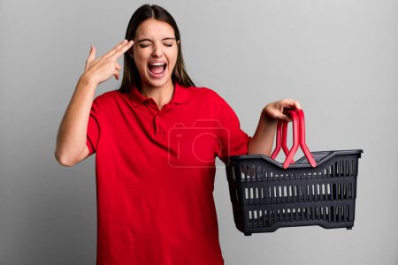 Photo for Young pretty woman looking unhappy and stressed, suicide gesture making gun sign. empty shopping basket concept - Royalty Free Image