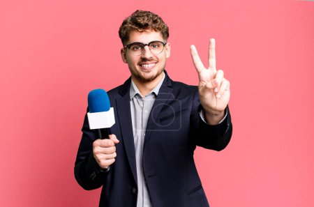 Foto de Joven hombre caucásico adulto sonriendo y mirando feliz, haciendo gestos de victoria o paz. periodista o presentador con micrófono - Imagen libre de derechos