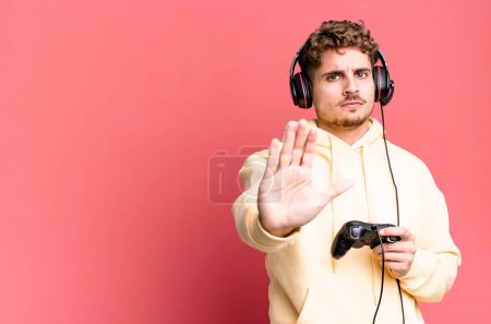 Foto de Joven hombre caucásico adulto que parece serio mostrando la palma abierta haciendo gesto de parada con auriculares y un controlador. concepto de jugador - Imagen libre de derechos