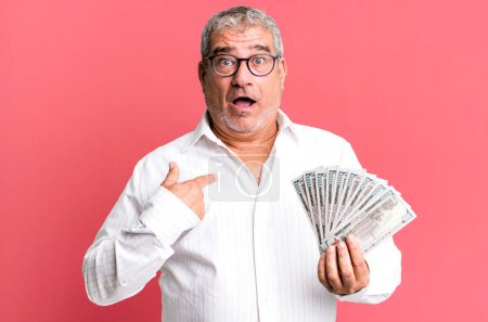Foto de Hombre mayor de mediana edad mirando sorprendido y sorprendido con la boca abierta, apuntando a sí mismo. concepto de billetes en dólares - Imagen libre de derechos