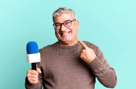 Téléchargez les photos : Homme âgé d'âge moyen souriant avec confiance pointant vers son propre sourire large. journaliste ou présentateur de télévision avec un micro - en image libre de droit