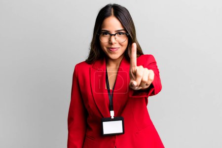 Photo for Hispanic pretty woman smiling proudly and confidently making number one with an accreditation card pass - Royalty Free Image