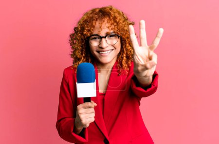 Foto de Cabello rojo bonita mujer sonriendo y buscando amigable, mostrando el número tres. concepto de periodista y presentador - Imagen libre de derechos