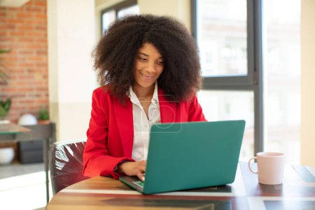 Téléchargez les photos : Jolie afro femme noire souriante et regardant avec une expression heureuse et confiante. femme d'affaires et ordinateur portable concept - en image libre de droit