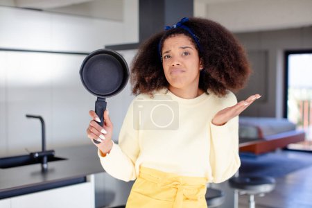 Foto de Mujer negra bastante afro encogiéndose de hombros, sintiéndose confundida e incierta. concepto de chef casero - Imagen libre de derechos
