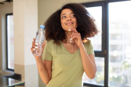 Foto de Bonita mujer negra afro sonriendo con una expresión feliz y segura con la mano en la barbilla. concepto de botella de agua - Imagen libre de derechos
