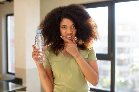 Téléchargez les photos : Jolie femme afro noire avec la bouche et les yeux grands ouverts et la main sur le menton. concept de bouteille d'eau - en image libre de droit
