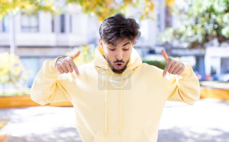 Photo for Young handsome man with open mouth pointing downwards with both hands, looking shocked, amazed and surprised - Royalty Free Image