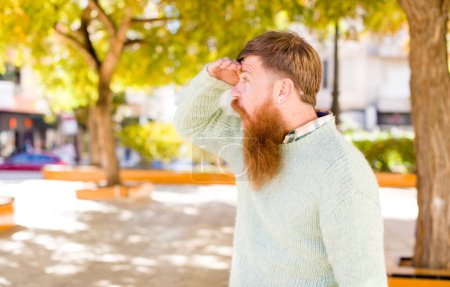 Photo for Red hair bearded man looking bewildered and astonished, with hand over forehead looking far away, watching or searching - Royalty Free Image