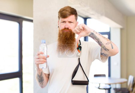 Foto de Hombre de pelo rojo sintiendo cruz, mostrando los pulgares hacia abajo con una botella de agua. concepto de fitness - Imagen libre de derechos