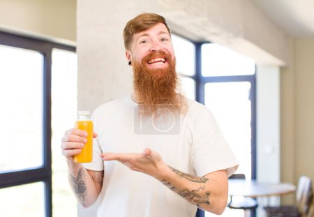 Foto de Hombre de pelo rojo sonriendo alegremente, sintiéndose feliz y mostrando un concepto con una botella de jugo de naranja - Imagen libre de derechos