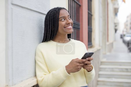 Foto de Mujer afro negro sonriendo con una expresión feliz y segura con la mano en la barbilla. usando un concepto de smartphone - Imagen libre de derechos