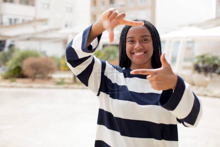 Foto de Afro bonita mujer negra sintiéndose feliz, amable y positiva, sonriendo y haciendo un retrato o marco de fotos con las manos - Imagen libre de derechos