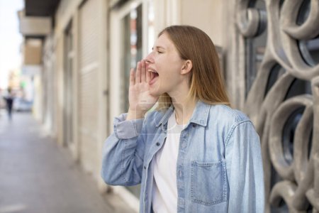 Photo for Pretty young woman profile view, looking happy and excited, shouting and calling to copy space on the side - Royalty Free Image