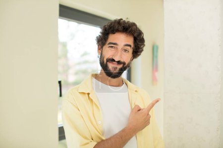 Téléchargez les photos : Jeune homme barbu fou souriant joyeusement, se sentant heureux et pointant vers le côté et vers le haut, montrant l'objet dans l'espace de copie - en image libre de droit