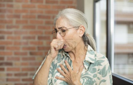Foto de Anciana bonita mujer sintiéndose enferma con dolor de garganta y síntomas de gripe, tos con la boca cubierta - Imagen libre de derechos
