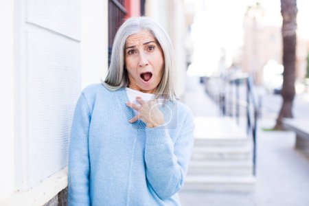 Photo for Senior retired pretty white hair woman feeling shocked, astonished and surprised, with hand on chest and open mouth, saying who, me? - Royalty Free Image