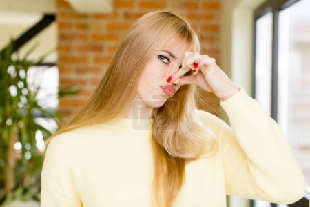 Téléchargez les photos : Jeune jolie femme se sentant dégoûtée, tenant le nez pour éviter de sentir une puanteur nauséabonde et désagréable. intérieur de la maison - en image libre de droit