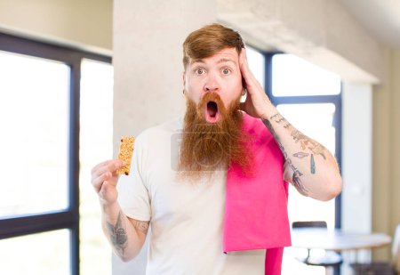 Photo for Red hair man feeling extremely shocked and surprised with a cereal bar. fitness concept - Royalty Free Image