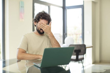 Photo for Young adult bearded man with a laptop covering eyes with one hand feeling scared or anxious, wondering or blindly waiting for a surprise - Royalty Free Image