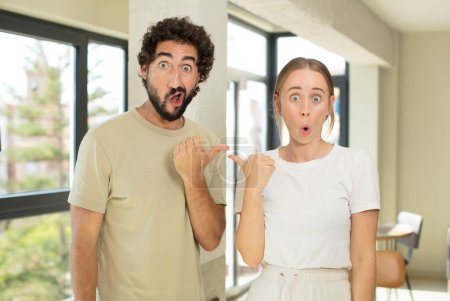 Photo for Young adult couple looking astonished in disbelief, pointing at object on the side and saying wow, unbelievable - Royalty Free Image
