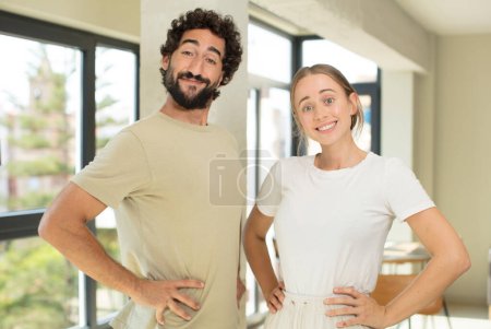 Foto de Joven pareja adulta sonriendo felizmente con las manos en la cadera y actitud confiada, orgullosa y amigable - Imagen libre de derechos