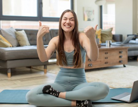 Foto de Mujer adulta joven practicando yoga sonriendo alegremente y luciendo feliz, sintiéndose despreocupada y positiva con ambos pulgares hacia arriba - Imagen libre de derechos