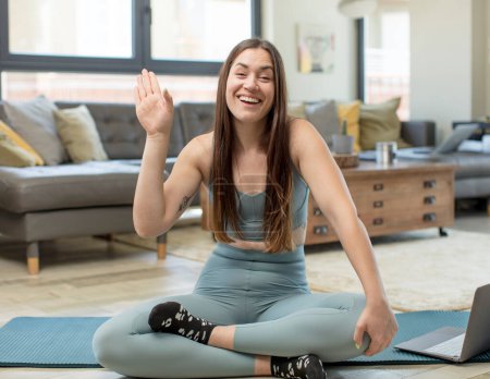 Foto de Mujer adulta joven practicando yoga sonriendo feliz y alegremente, saludándote con la mano, dándote la bienvenida y saludándote, o despidiéndote - Imagen libre de derechos