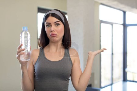 Téléchargez les photos : Jolie jeune femme haussant les épaules, se sentant confus et incertain. concept de bouteille d'eau - en image libre de droit