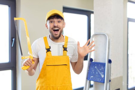 Photo for Young handsome man feeling happy and astonished at something unbelievable. handyman with a saw - Royalty Free Image