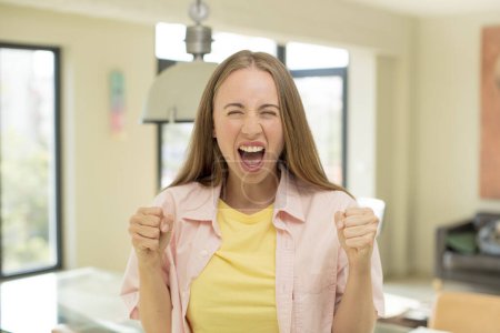 Photo for Pretty blond woman shouting aggressively with an angry expression or with fists clenched celebrating success - Royalty Free Image