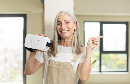 Photo for Pretty senior woman smiling cheerfully, feeling happy and pointing to the side. with vr goggles - Royalty Free Image