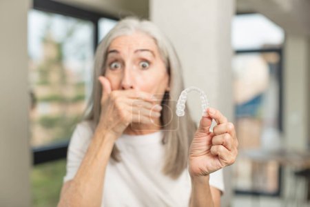 Photo for Pretty senior woman covering mouth with a hand and shocked or surprised expression. with a dental retainer - Royalty Free Image