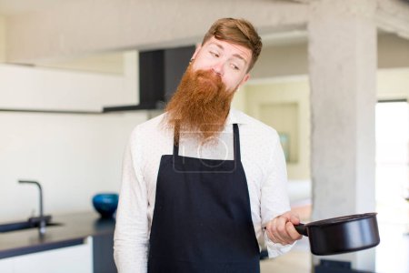 Téléchargez les photos : Homme aux cheveux roux souriant et regardant avec une expression heureuse et confiante dans une cuisine. concept de chef - en image libre de droit
